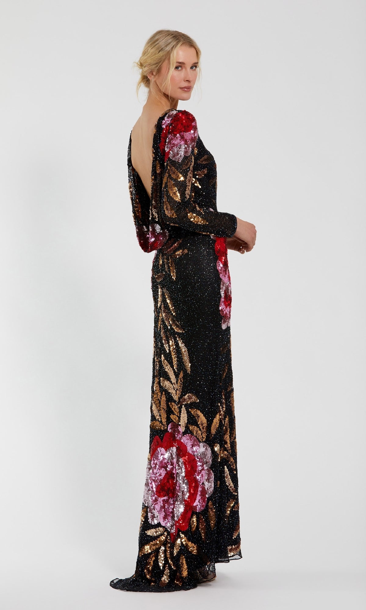 A woman stands sideways in the Mac Duggal 4566 long sleeve sequin formal gown, showcasing red, pink, and gold floral patterns on black fabric against a plain white background.
